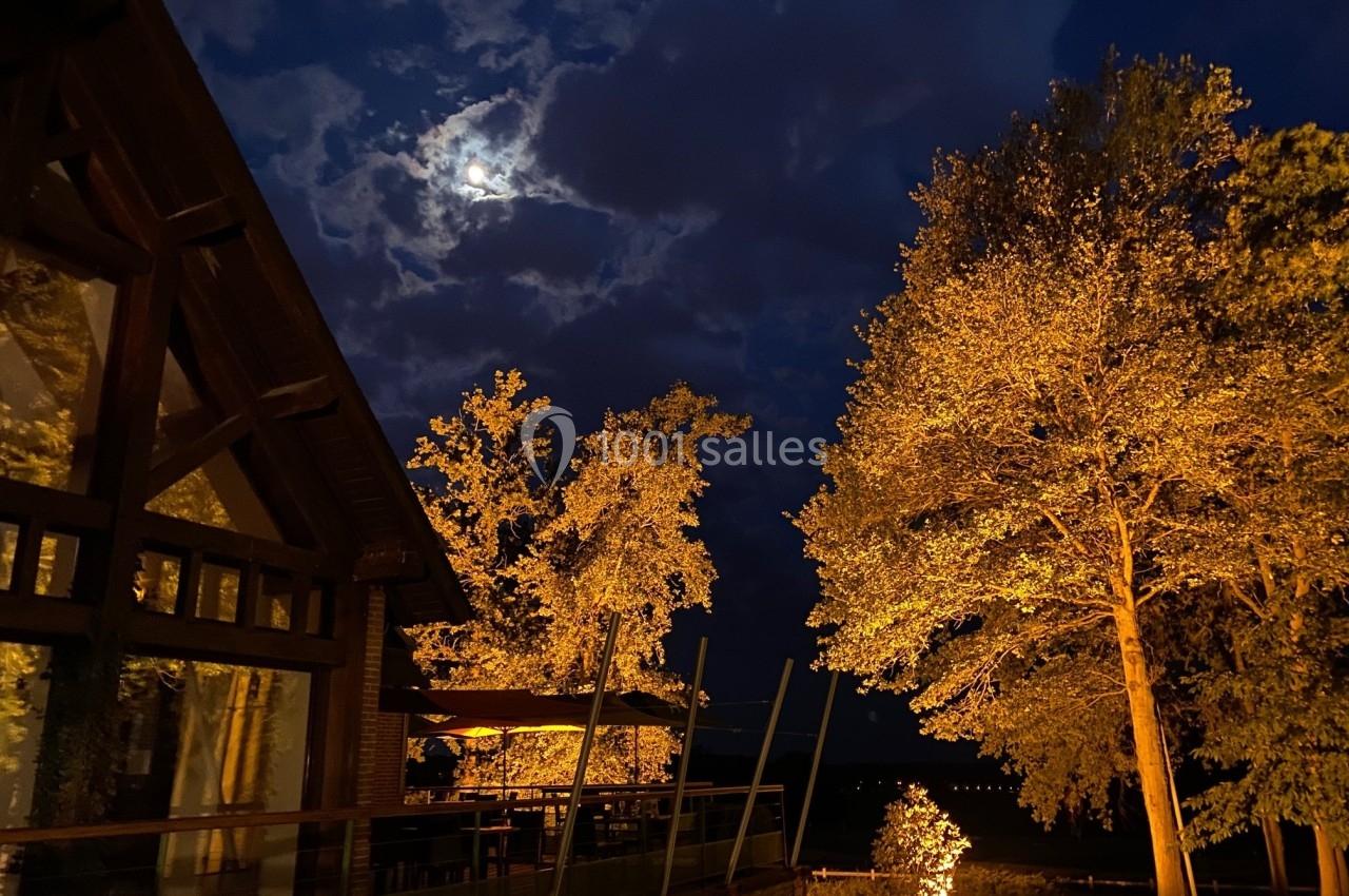 Vue nocturne d'une maison en bois éclairée, entourée d'arbres illuminés, sous un ciel nuageux avec la lune visible.