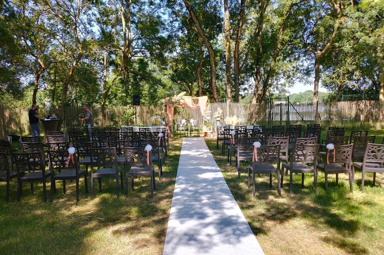 Allée centrale bordée de chaises noires menant à une arche décorée pour une cérémonie en plein air sous des arbres.