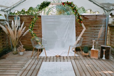 Espace extérieur décoré avec arche florale, deux chaises transparentes et fond blanc sous une tente.