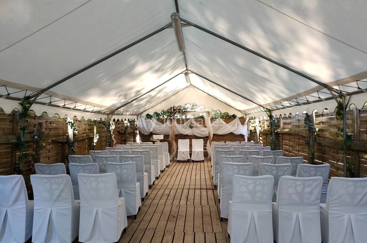 Salle de cérémonie en plein air sous une tente, avec des chaises blanches alignées et une décoration en bois et tissu.