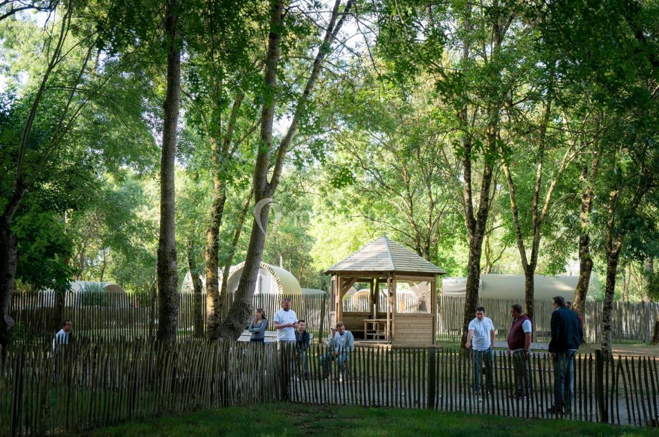 Un groupe de personnes discute près d'un kiosque en bois entouré d'arbres et de structures légères dans un espace naturel.