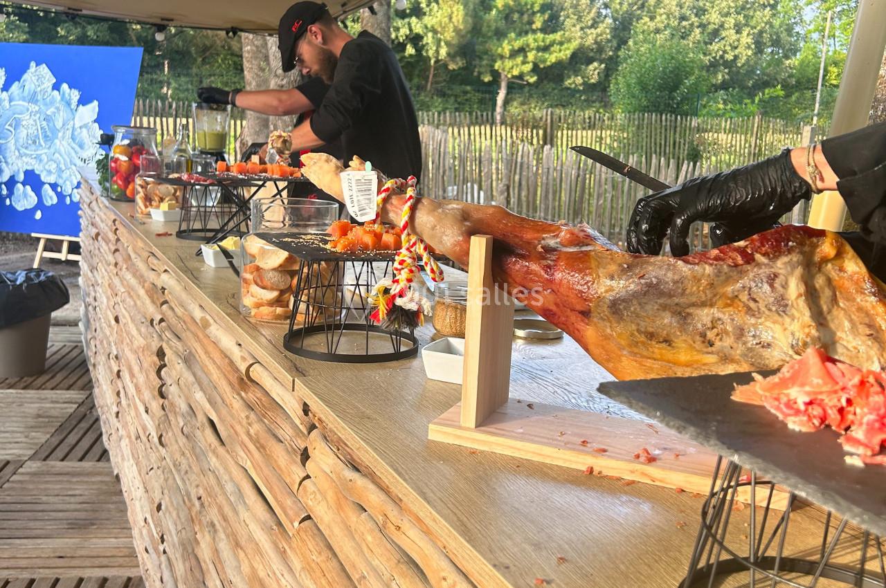 Des cuisiniers découpent des jambons sur un comptoir en bois lors d'un événement en plein air.