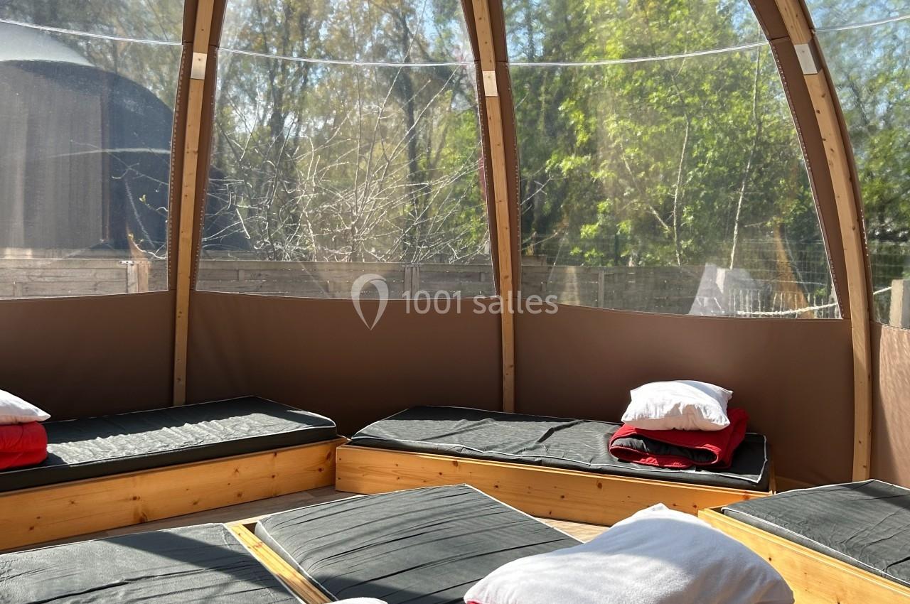 Intérieur d'une structure en bois avec des matelas et des couvertures rouges, entourée de grandes baies vitrées donnant sur…