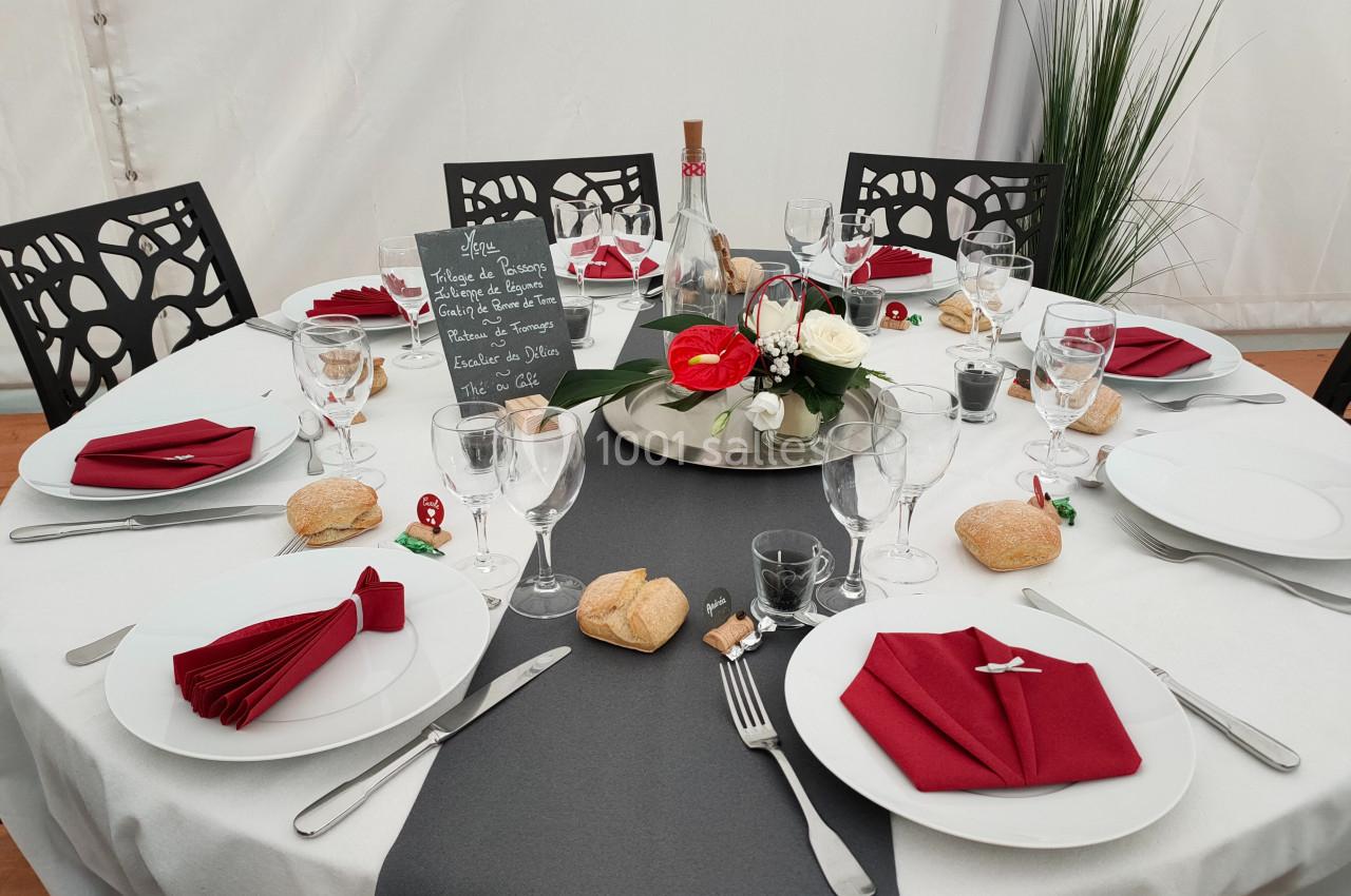 Table dressée pour un repas, avec nappes blanches et grises, serviettes rouges pliées, vaisselle, pain et décoration florale.