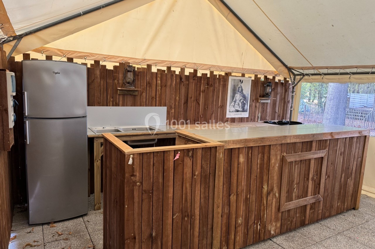Cuisine extérieure sous une tente avec un comptoir en bois, un évier, un réfrigérateur et un mur en bois décoré.