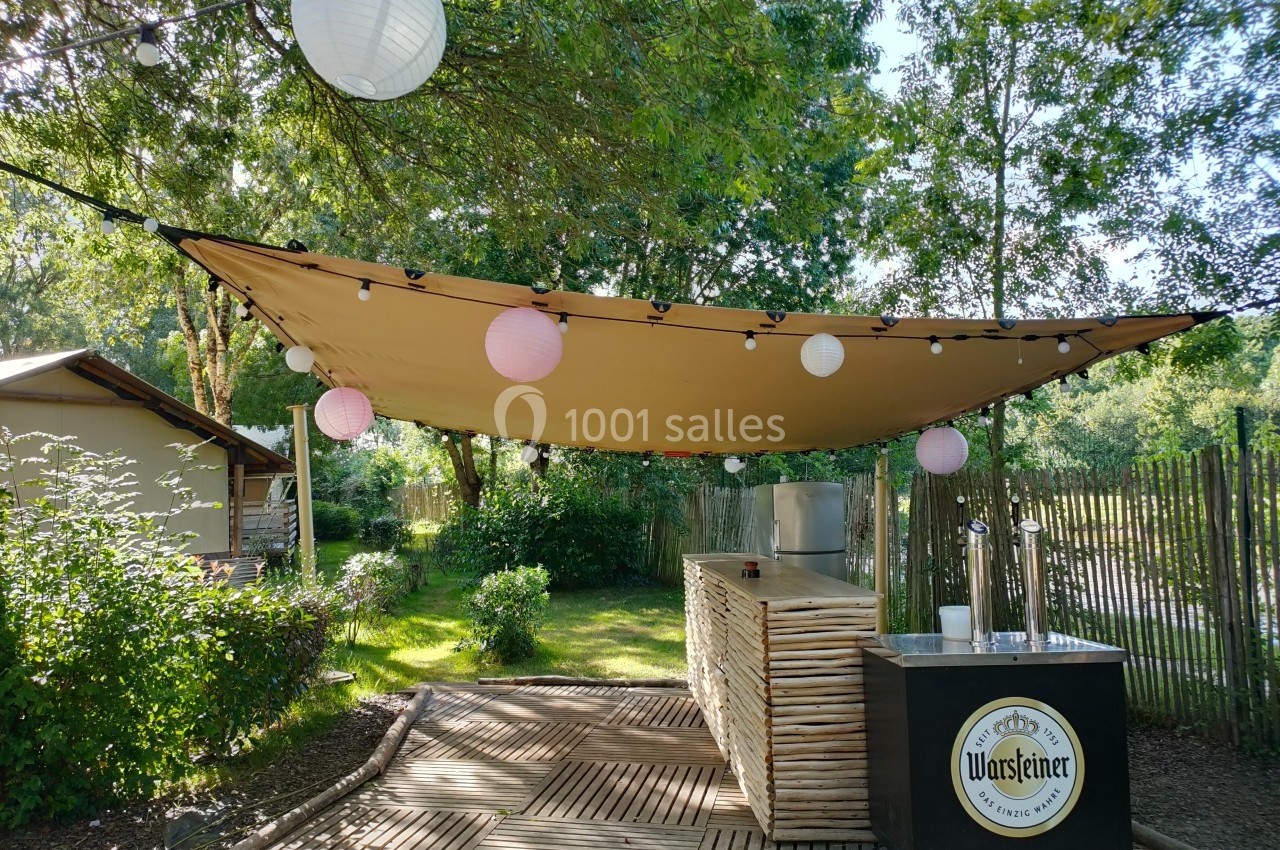 Bar extérieur en bois sous une toile tendue, décoré de lampions et entouré de végétation.