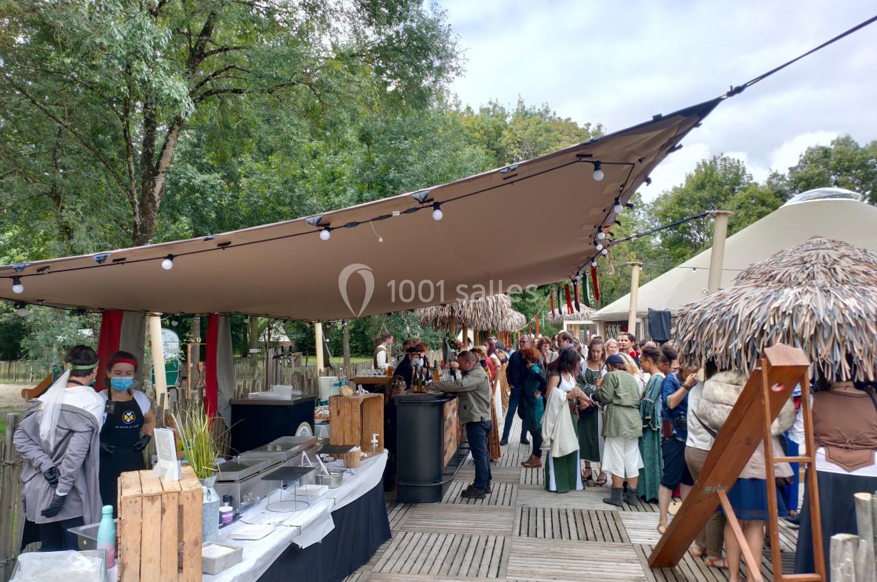File d'attente devant un stand de restauration en plein air, sous une grande toile tendue, entouré d'arbres.