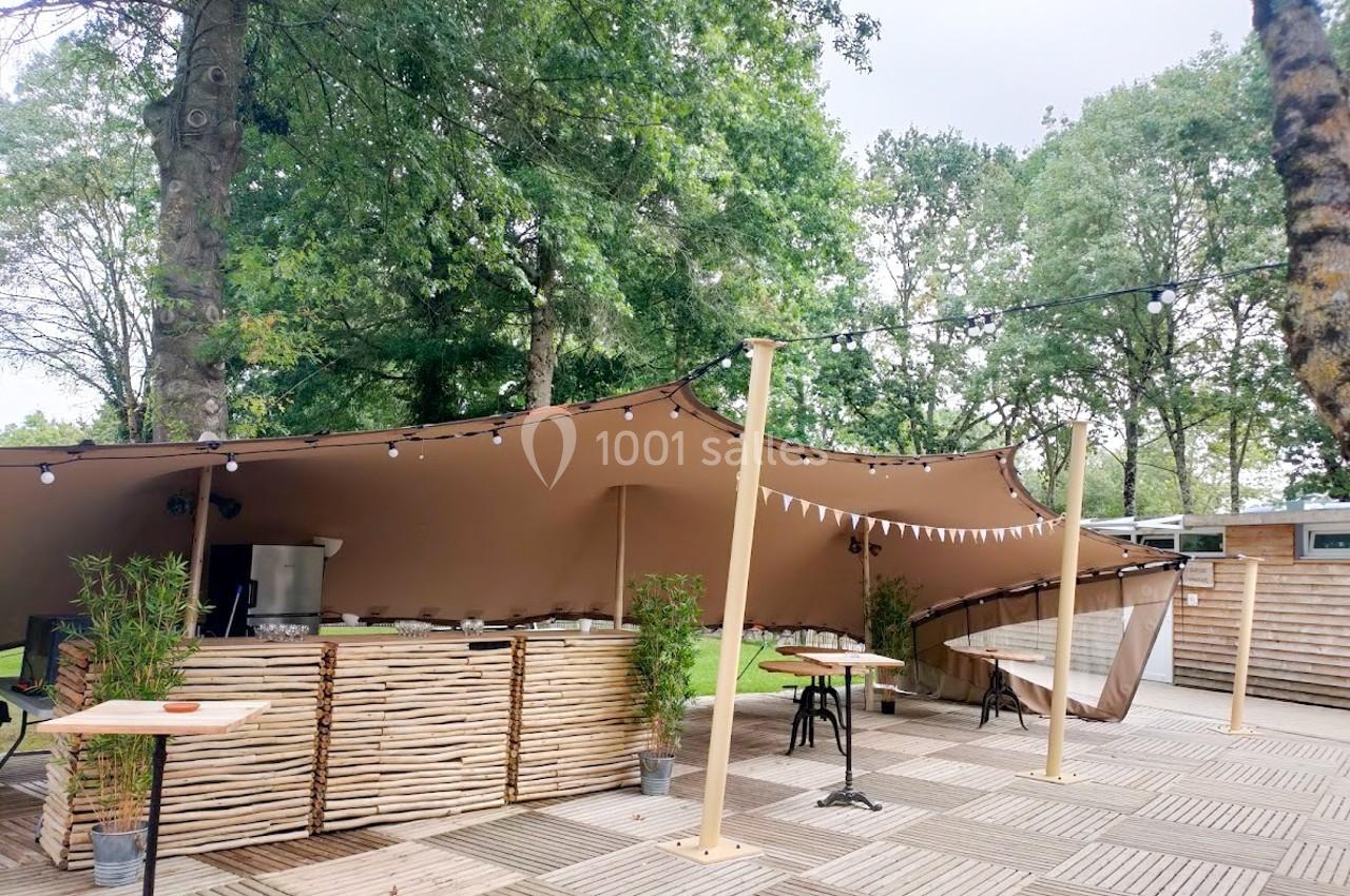 Terrasse extérieure avec tente beige, tables hautes, plantes en pot et guirlandes lumineuses, entourée d'arbres.