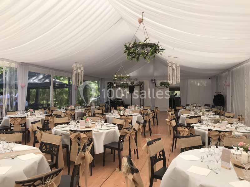 Salle de réception décorée avec des tables rondes dressées, des chaises ornées de nœuds et des suspensions florales.