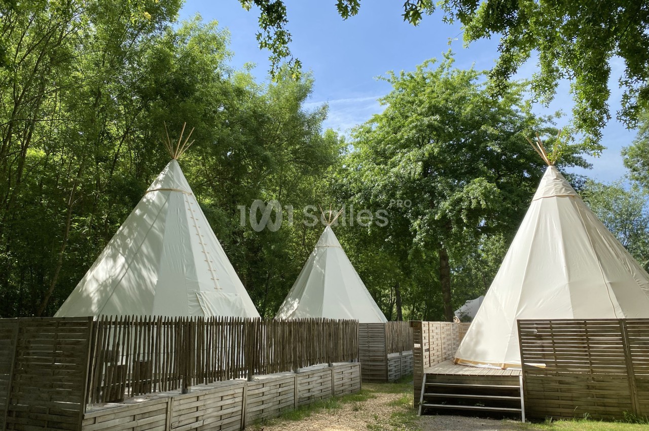 Trois tipis blancs entourés de clôtures en bois, installés dans une clairière verdoyante sous un ciel dégagé.