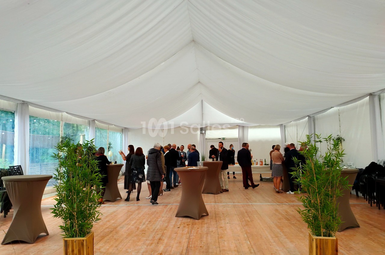 Personnes rassemblées dans une grande tente blanche avec des tables hautes et des plantes en premier plan.