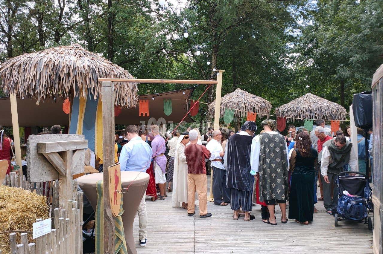 Groupe de personnes en costumes médiévaux rassemblées sous des abris en paille dans un cadre extérieur boisé.