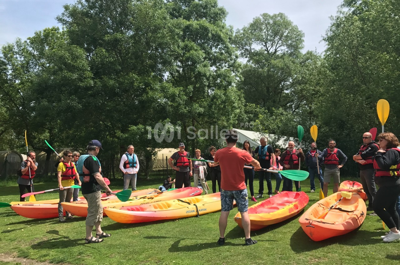 Un groupe de personnes en gilets de sauvetage écoute des instructions près de kayaks sur une pelouse verdoyante.