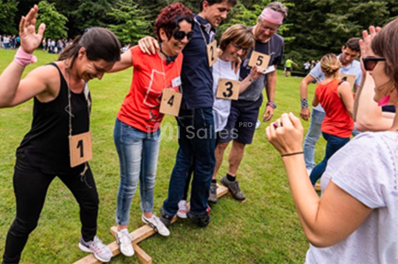 Des personnes en équipe participent à une activité collaborative sur une pelouse, marchant ensemble sur des planches en bois.