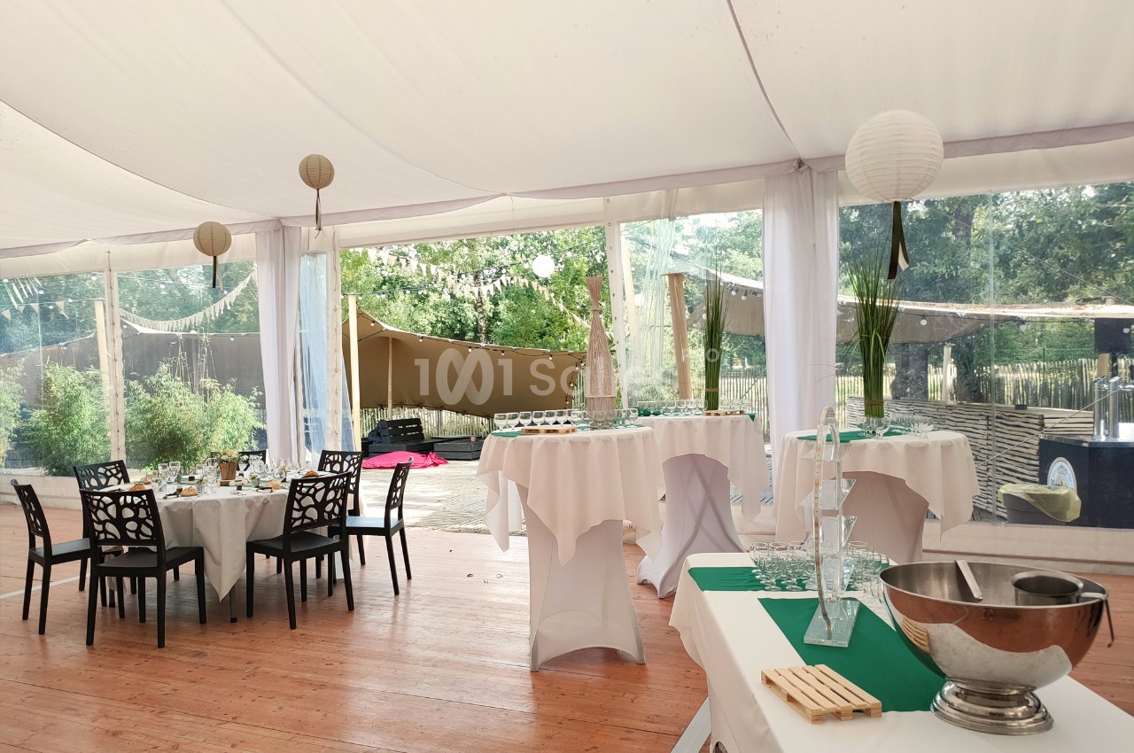 Salle de réception sous une tente avec tables dressées, nappes blanches et vue sur un espace extérieur arboré.