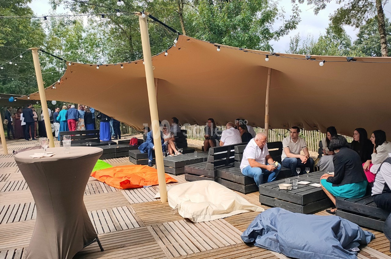 Personnes assises sous une tente beige sur une terrasse en bois, discutant autour de tables basses avec des poufs colorés.