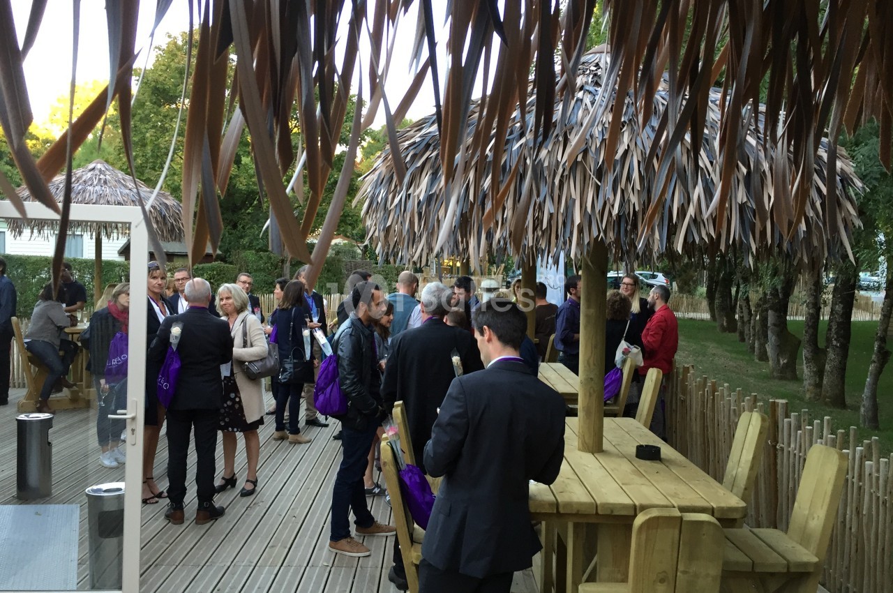 Groupe de personnes discutant sur une terrasse en bois, entourée de parasols en feuilles et d'arbres.