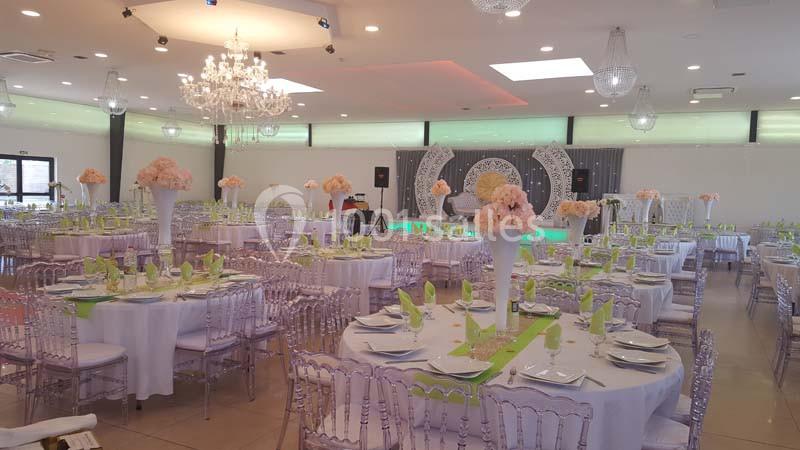 Salle de réception décorée avec des tables rondes, nappes blanches, chaises transparentes et centres de table fleuris.