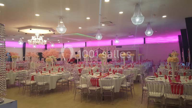 Salle de réception décorée avec des tables dressées, des chaises transparentes et des éclairages roses et blancs.