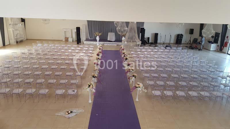 Salle de réception aménagée pour une cérémonie, avec allée centrale violette bordée de fleurs et chaises transparentes.