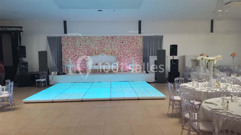 Salle de réception décorée avec une scène ornée de fleurs, un sol lumineux et des tables dressées pour un événement.