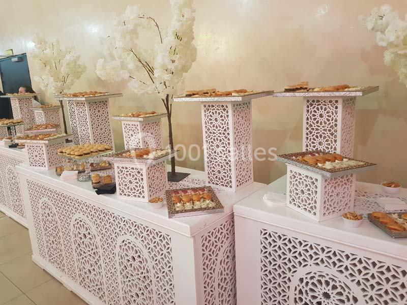 Buffet de pâtisseries orientales disposées sur des présentoirs blancs ornés de motifs ajourés, avec décor floral blanc.