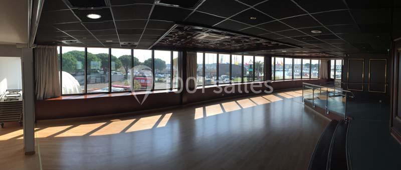 Salle spacieuse avec parquet, grandes baies vitrées donnant sur un port et plafond sombre avec éclairage intégré.