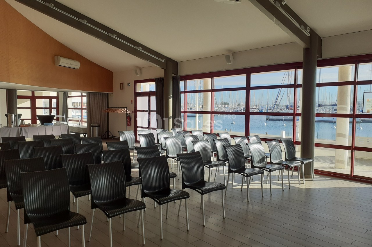 Salle lumineuse avec des rangées de chaises noires, grandes baies vitrées donnant sur un port de plaisance.