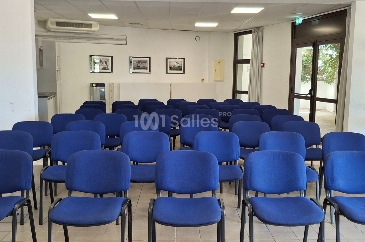 Salle de réunion avec des rangées de chaises bleues vides, éclairée par la lumière naturelle provenant des fenêtres.