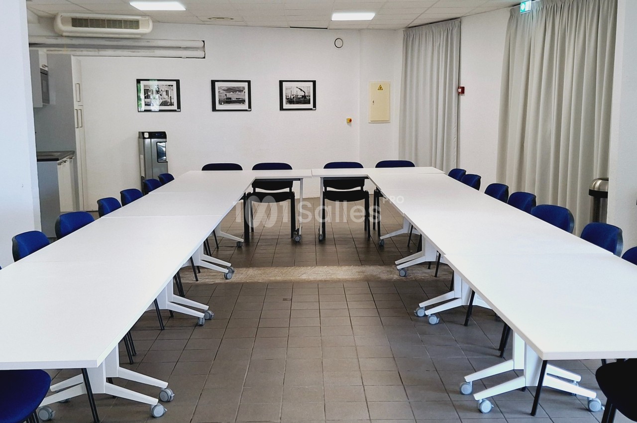 Salle de réunion lumineuse avec tables blanches disposées en U, chaises bleues et cadres accrochés au mur.