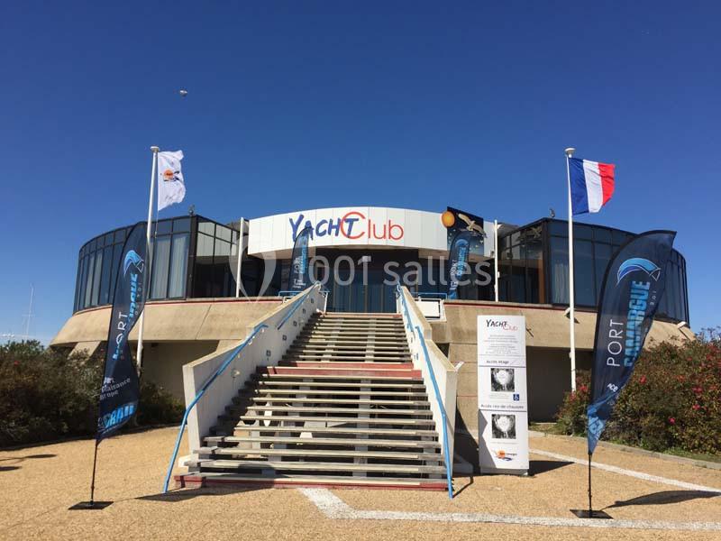 Entrée principale d'un bâtiment circulaire marqué ’Yacht Club’, avec des drapeaux et des bannières devant les escaliers.