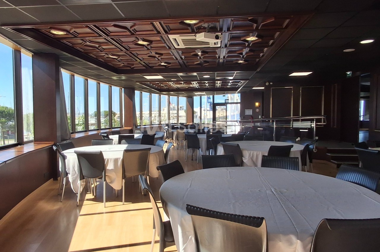 Salle lumineuse avec de grandes baies vitrées, tables rondes dressées et chaises noires, vue sur un extérieur arboré.