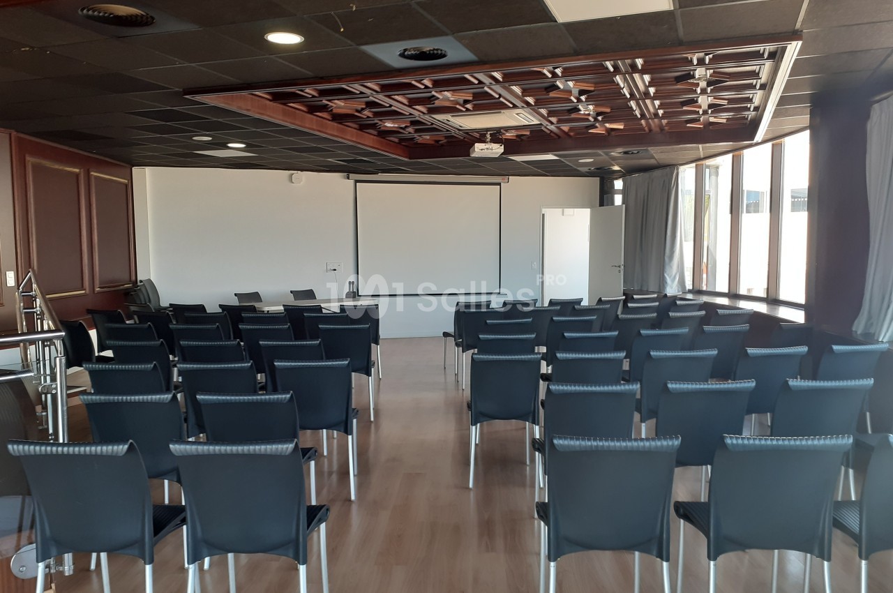 Salle de conférence vide avec des rangées de chaises noires, un écran de projection et un plafond décoratif en bois.