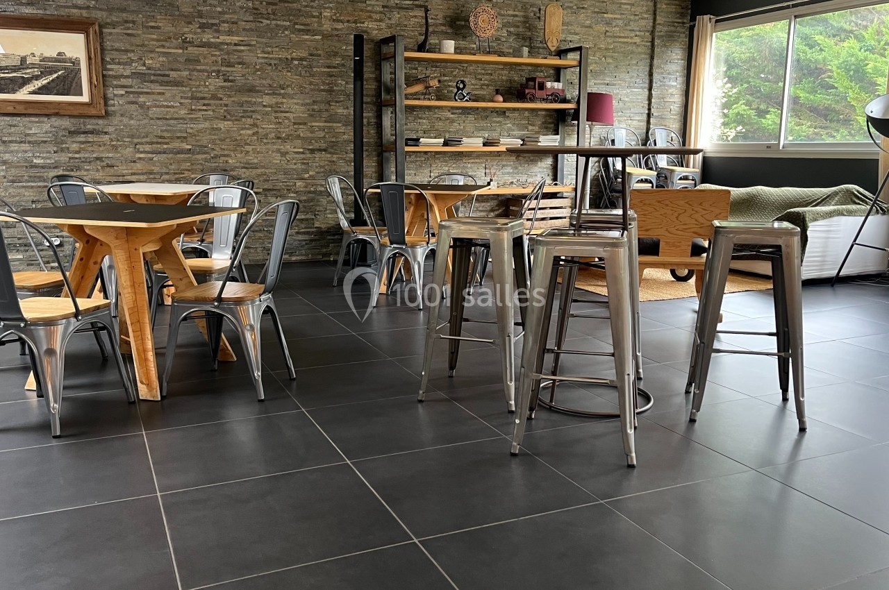 Salle lumineuse avec tables en bois, chaises métalliques, étagères décoratives et mur en pierre apparente.