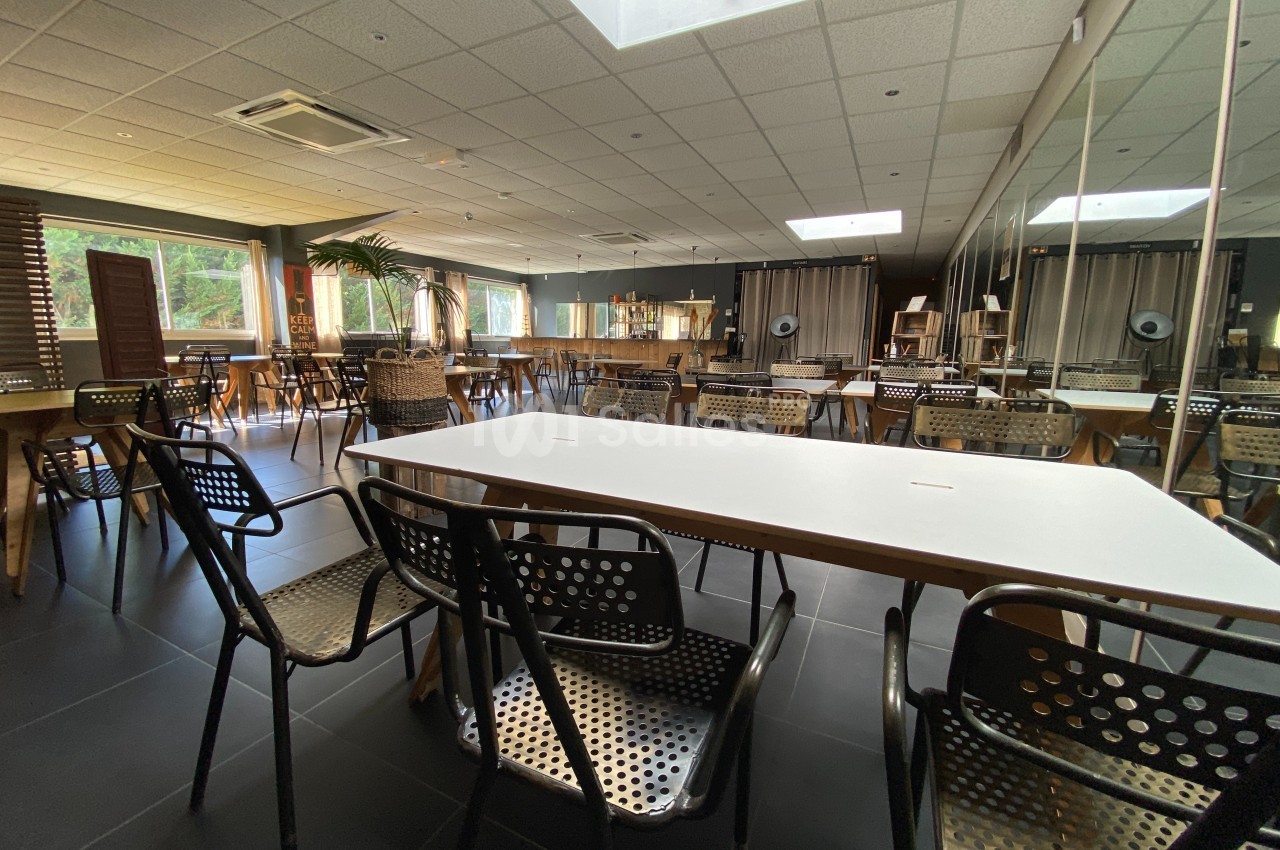 Salle lumineuse avec tables blanches, chaises en métal ajouré, grandes fenêtres et miroirs sur un mur.