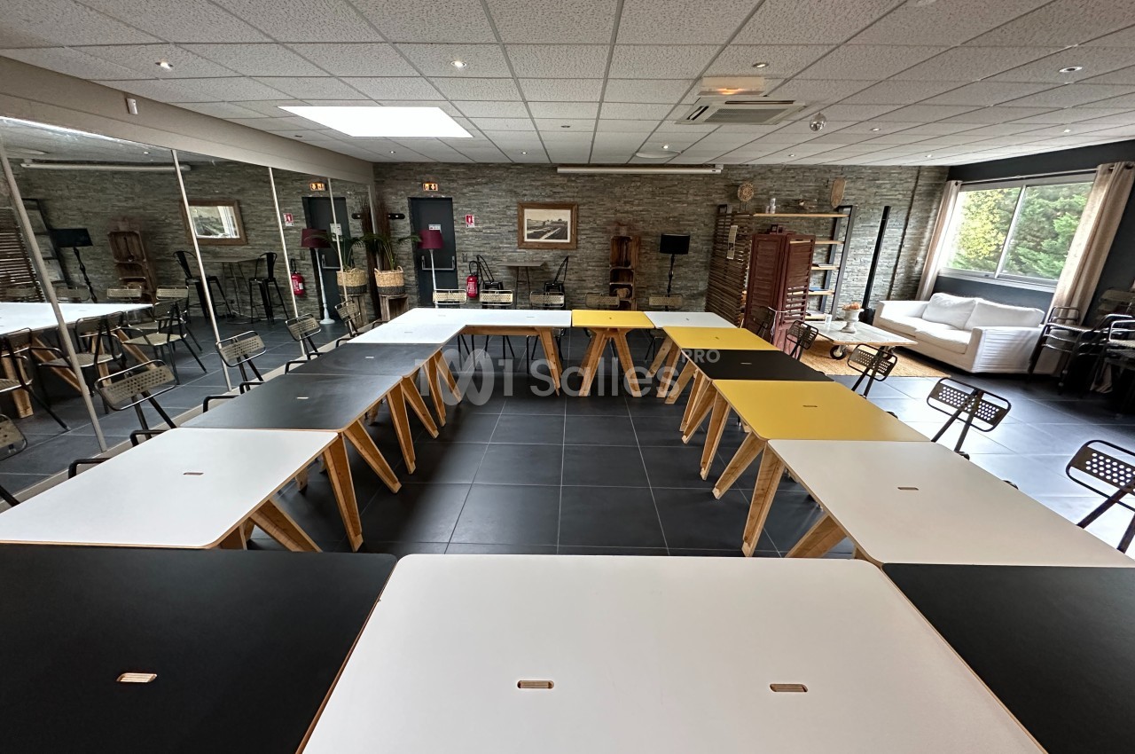 Salle de réunion avec des tables disposées en U, chaises noires, murs en pierre et grandes fenêtres laissant entrer la…