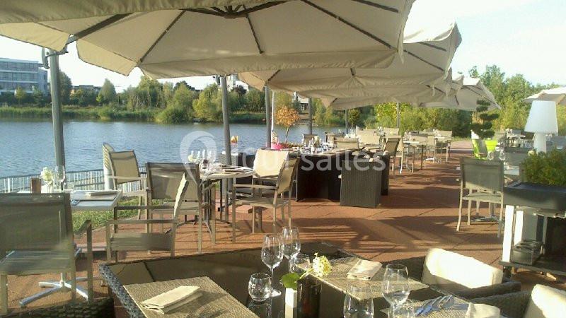 Terrasse de restaurant avec tables dressées, chaises et parasols, offrant une vue sur un lac et un paysage verdoyant.
