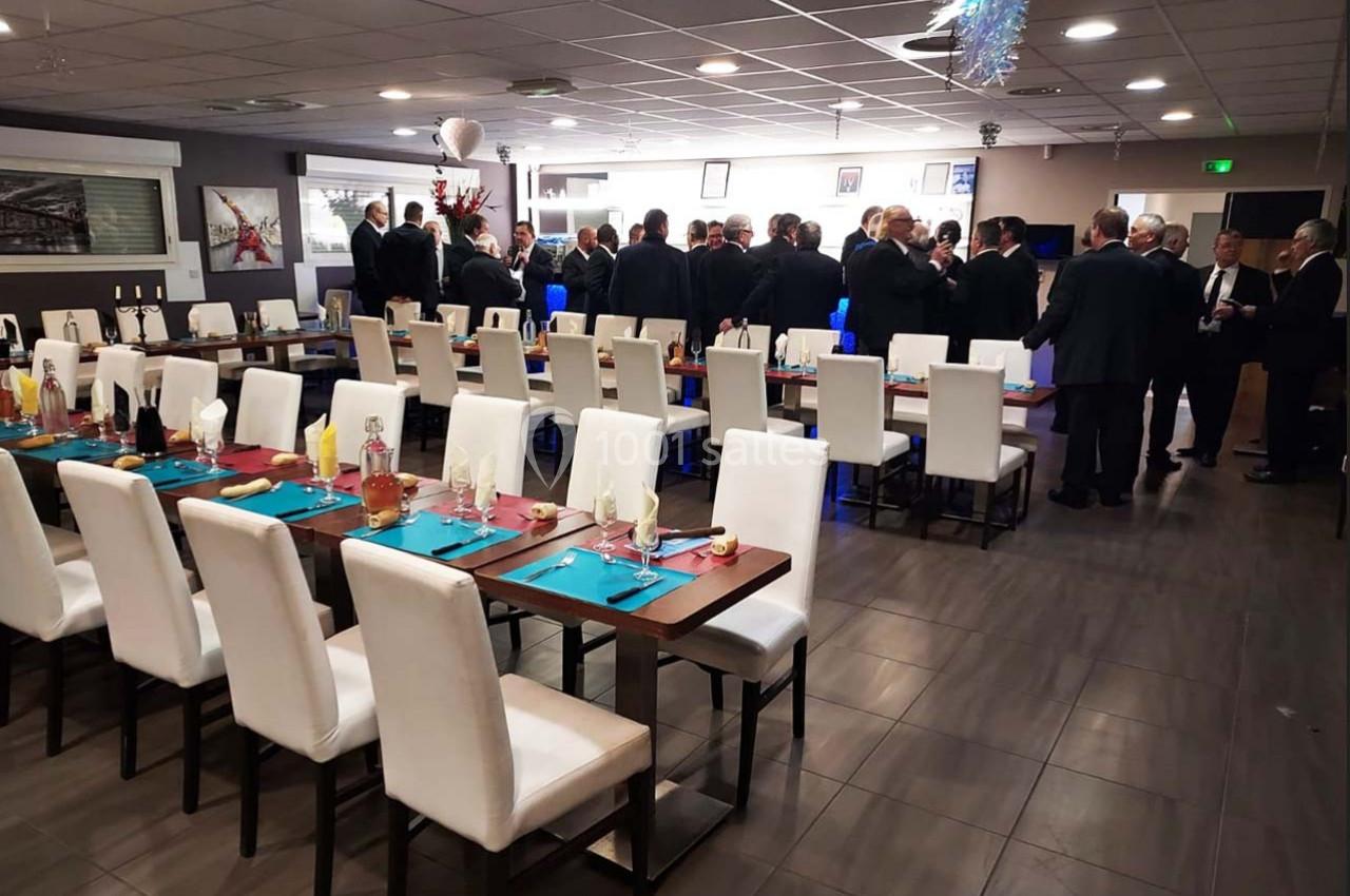Salle de réception avec tables dressées et invités en costume debout près d'un buffet ou d'un bar.