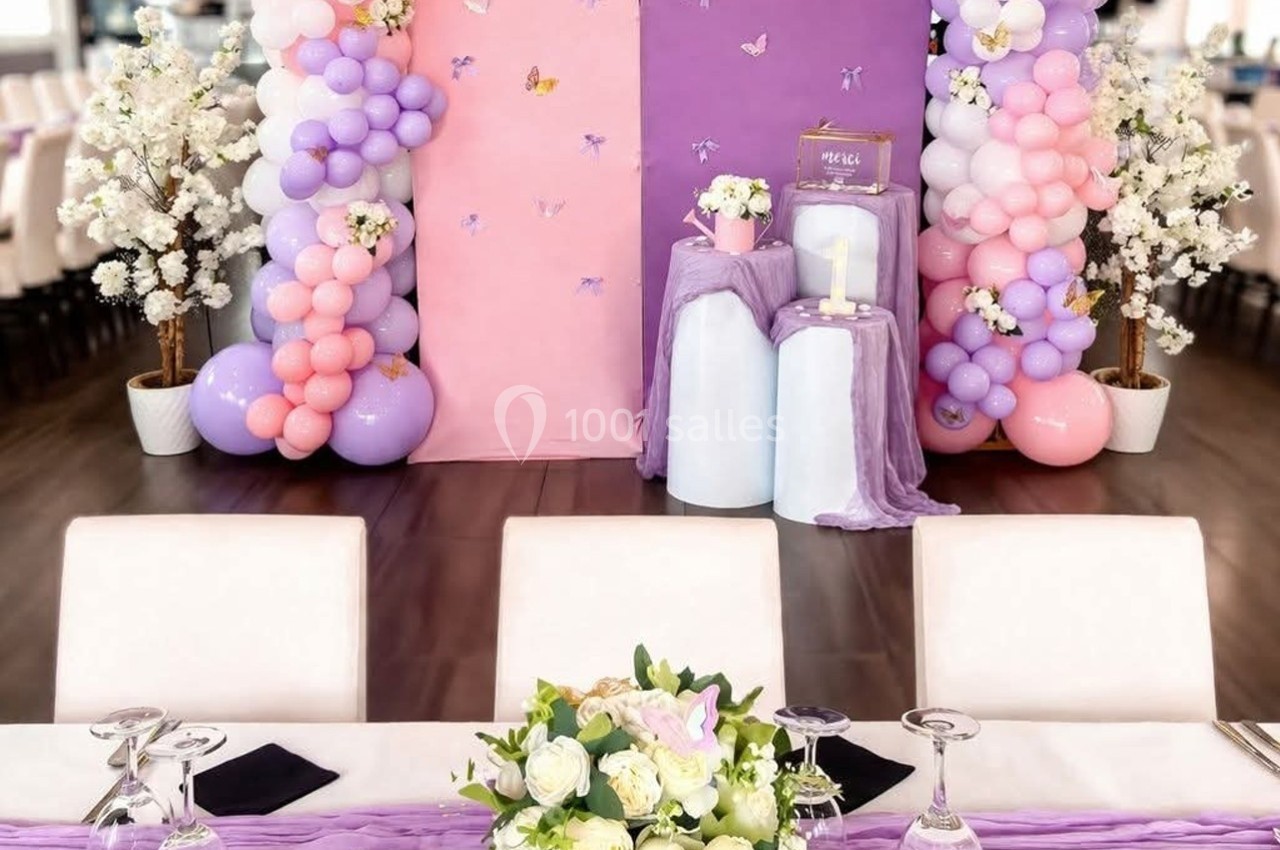 Décoration de salle avec ballons pastel, fleurs et bougies, vue depuis une table dressée avec nappe violette.