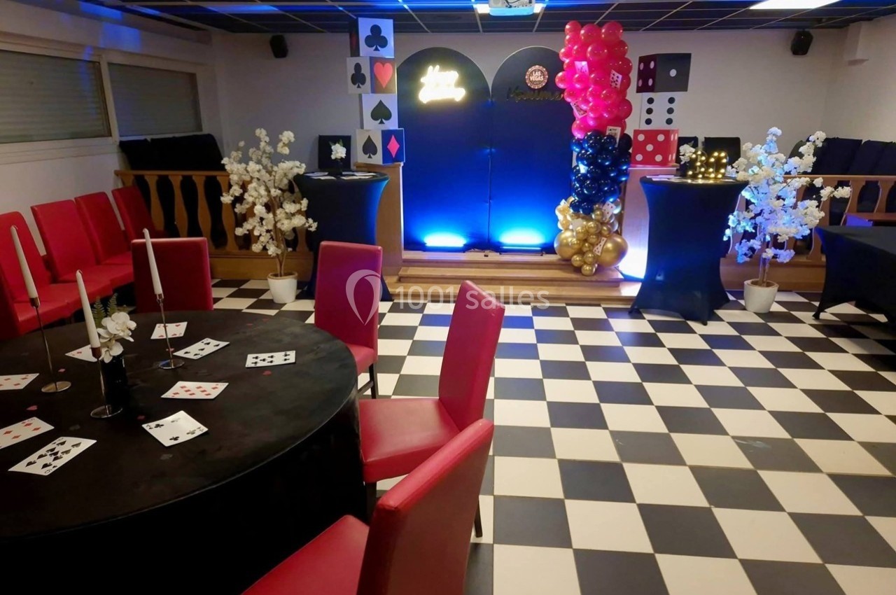 Salle décorée sur le thème du casino avec tables, chaises rouges, ballons et cartes à jouer sur un sol en damier.