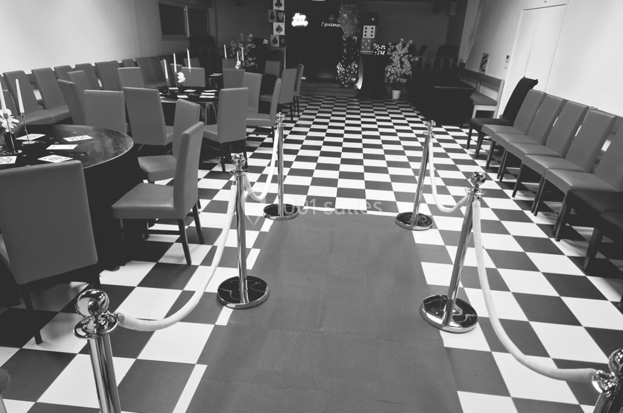 Salle avec sol à carreaux noirs et blancs, rangées de chaises, tables et cordons délimitant un tapis central.