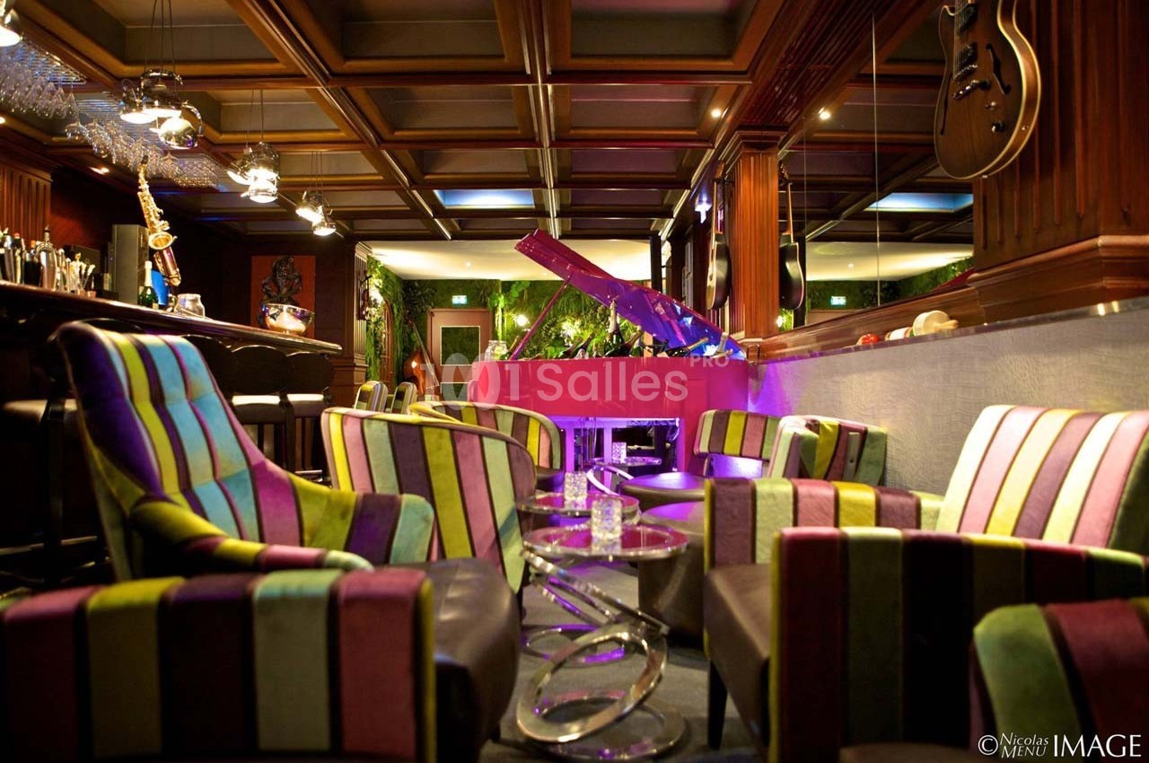 Salon avec fauteuils colorés, piano rose, bar en arrière-plan et décorations modernes dans une ambiance chaleureuse.