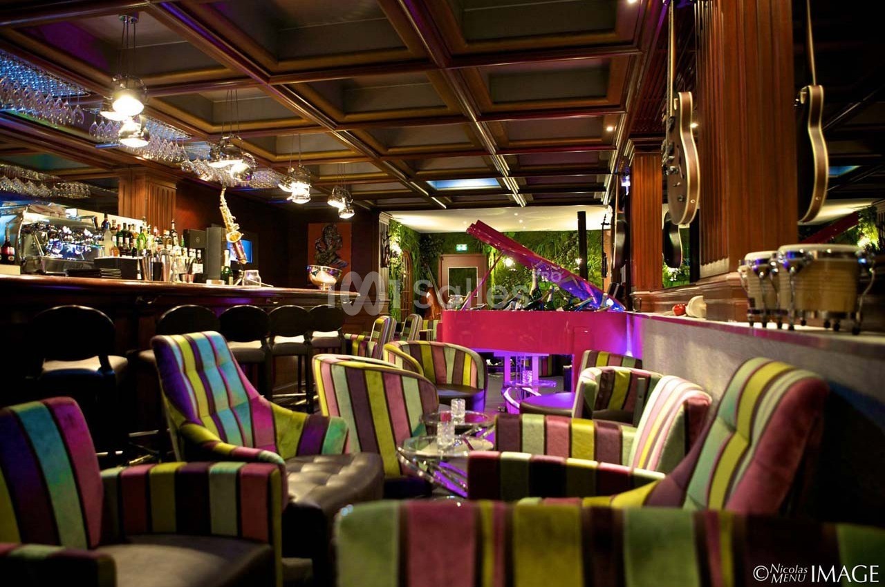 Salon coloré avec fauteuils rayés, bar en arrière-plan et piano rose éclairé dans une ambiance tamisée.