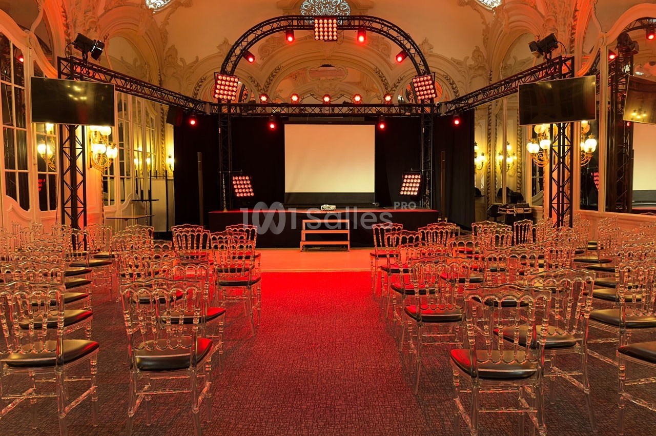 Salle de conférence élégante avec des chaises transparentes, une scène équipée de projecteurs rouges et un écran blanc.