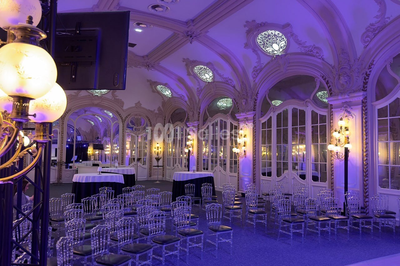 Salle élégante avec des chaises transparentes, tables hautes et éclairage violet, ornée de moulures et de miroirs.