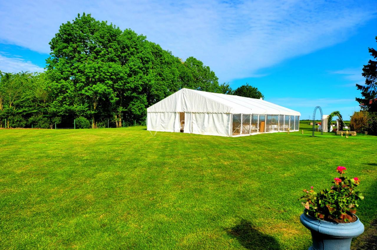 Tente blanche installée sur une pelouse verte, entourée d'arbres sous un ciel bleu dégagé.