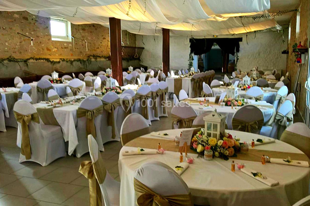 Salle décorée pour un événement, avec des tables rondes dressées, nappes blanches et chaises ornées de nœuds dorés.