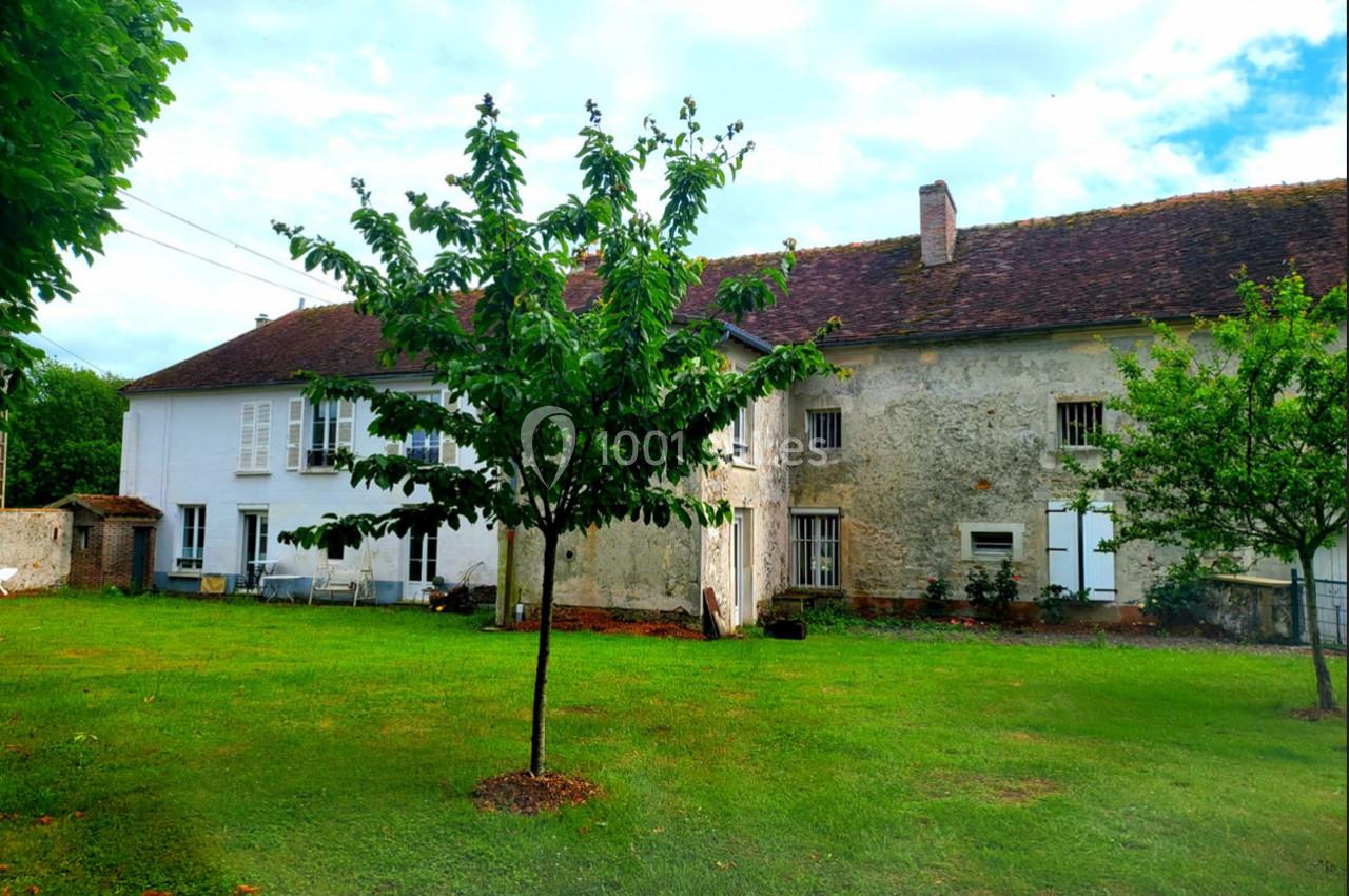 Façade d'une maison ancienne avec un jardin verdoyant, un arbre au premier plan et un ciel légèrement nuageux.
