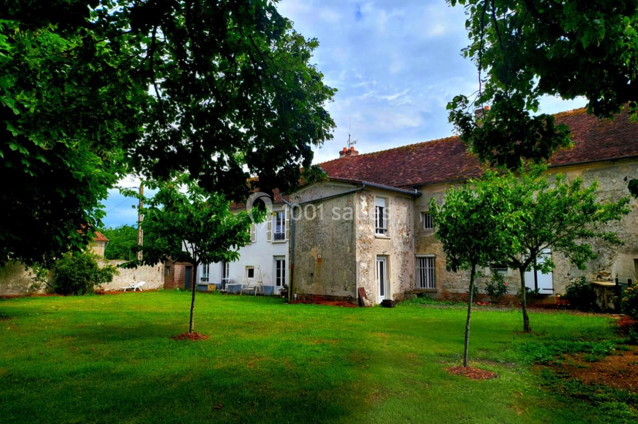Grande maison en pierre avec toit en tuiles, entourée d'un jardin verdoyant et d'arbres sous un ciel légèrement nuageux.