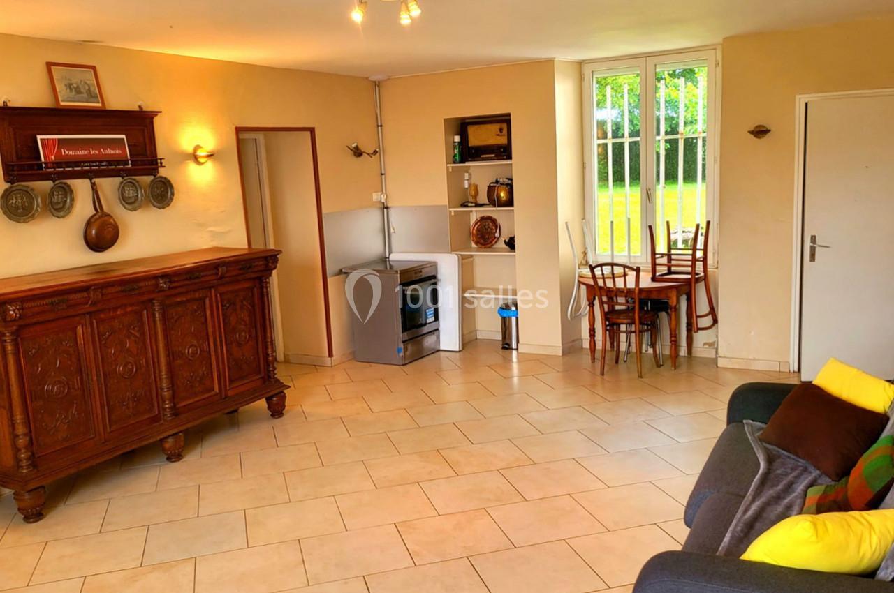 Salon lumineux avec carrelage beige, canapé, meuble ancien, coin cuisine équipé et table près d'une fenêtre.